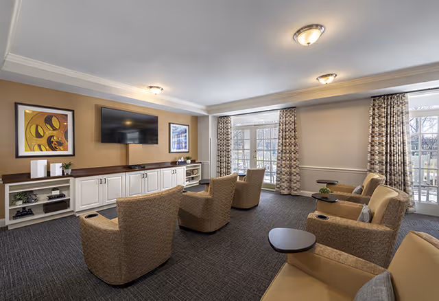 A bright common living room with cushioned armchairs arranged toward a wall-mounted TV above built-in cabinets and large glass doors with patterned curtains.