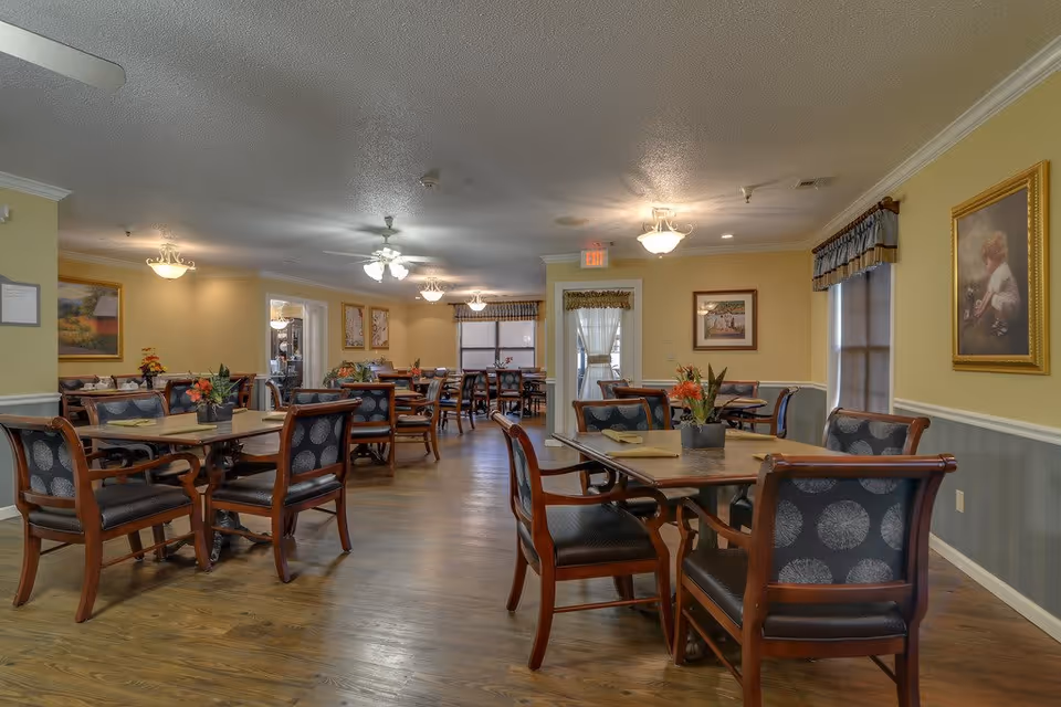 Spacious senior living dining room with multiple wooden tables and upholstered chairs and floral centerpieces under ceiling lights.
