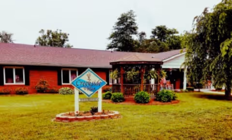 Exterior view of Creekside Villas Retirement facility showing a single-story brick building with a red roof, a green lawn, a wooden gazebo with hanging plants, and a sign in front that reads 'Creekside Villas'. Trees are visible in the background.