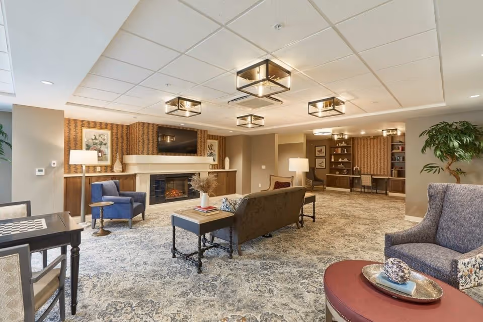 A spacious and well-lit living room area in a senior living facility featuring a patterned carpet, multiple seating options including armchairs and a sofa, a fireplace with a TV mounted above it, decorative lamps, framed artwork on the walls, and built-in shelves with a desk area in the background.