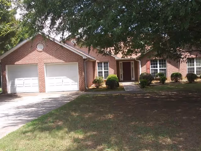 Front exterior view of a single-story brick house with a two-car garage, a concrete driveway, and a front entrance pathway. The house is surrounded by a lawn with some bushes and large trees providing shade.