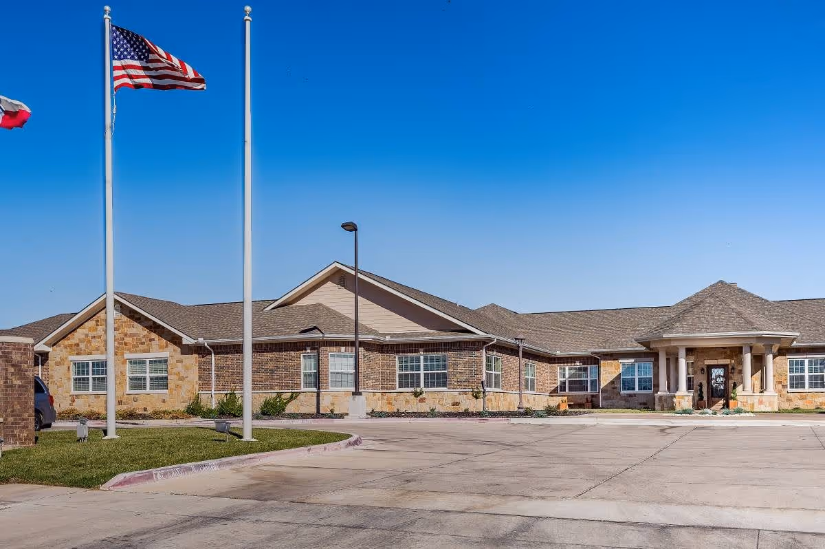 Exterior view of a single-story brick and stone building under a clear blue sky with two flagpoles displaying the American flag and another flag. The building has multiple windows and a covered entrance with columns. There is a paved driveway and some landscaping with grass and small plants.