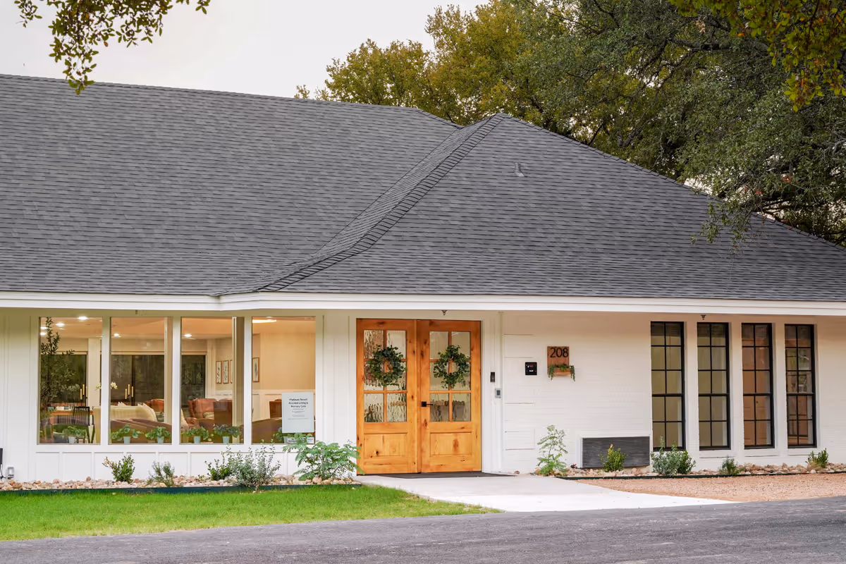 Front entrance of a single-story assisted living building with large windows, double wooden doors adorned with wreaths, and a small landscaped lawn.