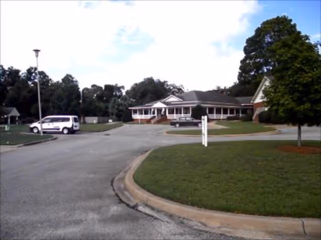 View of a single-story building with a wrap-around porch, surrounded by a driveway and green lawn with trees. Two vehicles are parked near the building under a partly cloudy sky.
