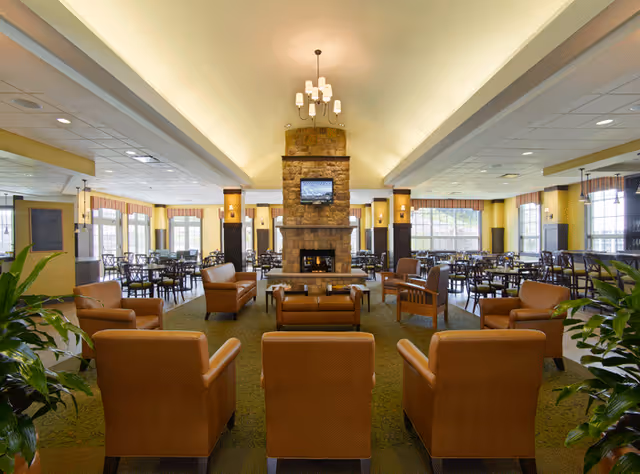 A spacious and well-lit common area in a senior living facility featuring a central stone fireplace with a mounted TV above it. The room is furnished with multiple brown leather armchairs arranged around the fireplace, and several dining tables and chairs are visible in the background near large windows that allow natural light to fill the space. Green plants are placed on either side of the seating area, and a chandelier hangs from the ceiling.