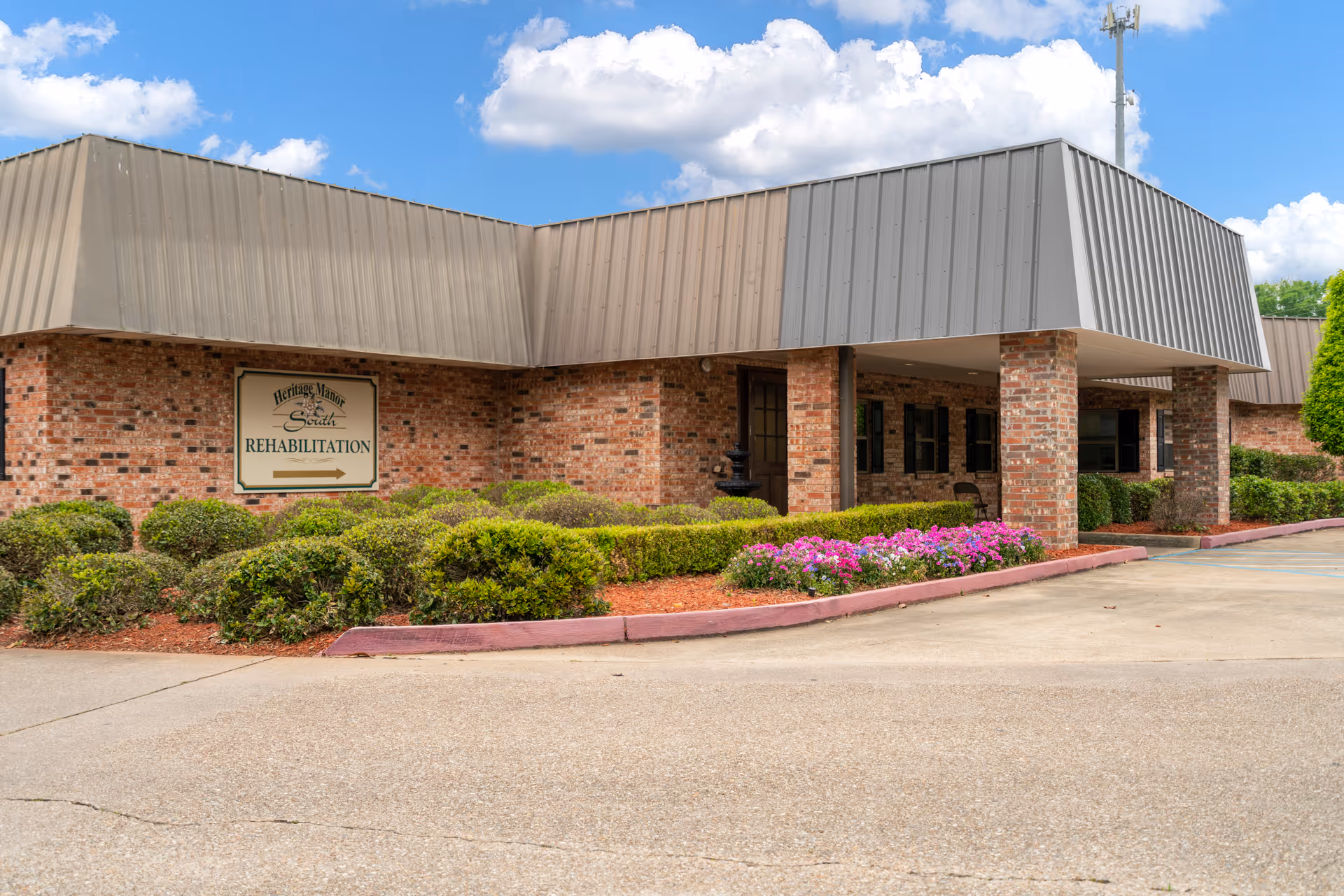 Exterior view of Heritage Manor South building with brick walls, a covered entrance supported by brick columns, landscaped bushes and flowers, and a sign indicating Rehabilitation services.