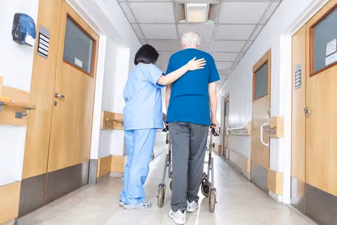 A healthcare worker in blue scrubs is assisting an elderly man using a walker as they walk down a hallway in a care facility.