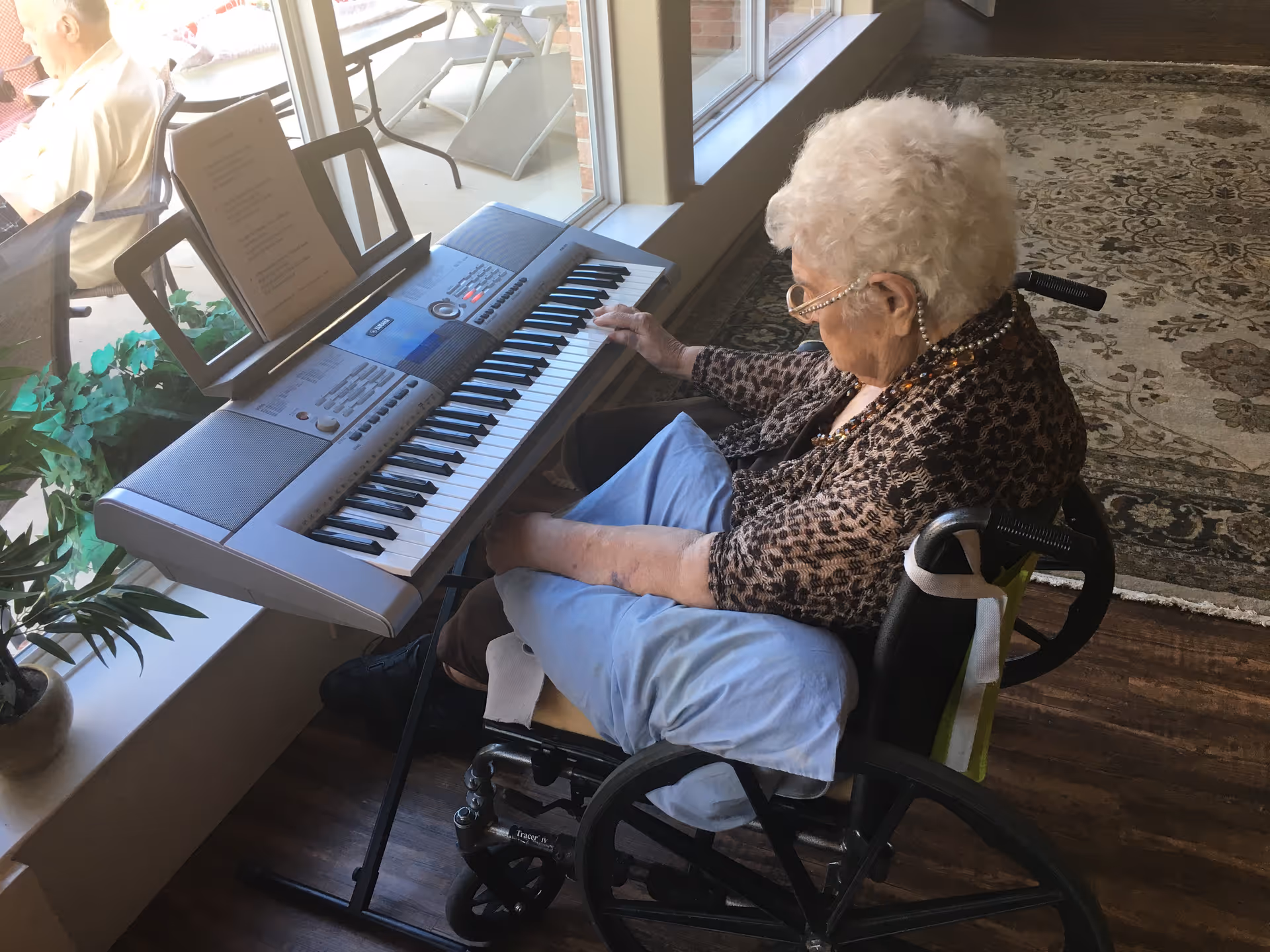 An elderly woman in a wheelchair playing an electronic keyboard placed near a large window. She is wearing glasses and a patterned top, with a blue pillow on her lap. Outside the window, another elderly person is sitting on a chair on a patio. The room has wooden floors and a patterned rug.