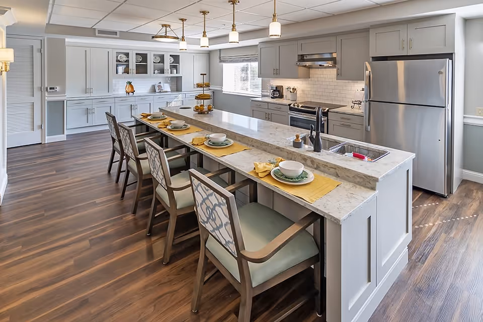 A modern kitchen with a long marble island countertop featuring four place settings with bowls, plates, and yellow placemats. The kitchen has light gray cabinets, stainless steel appliances including a refrigerator and stove, a sink on the island, and a window with blinds letting in natural light. The floor is wood with a rich brown finish.