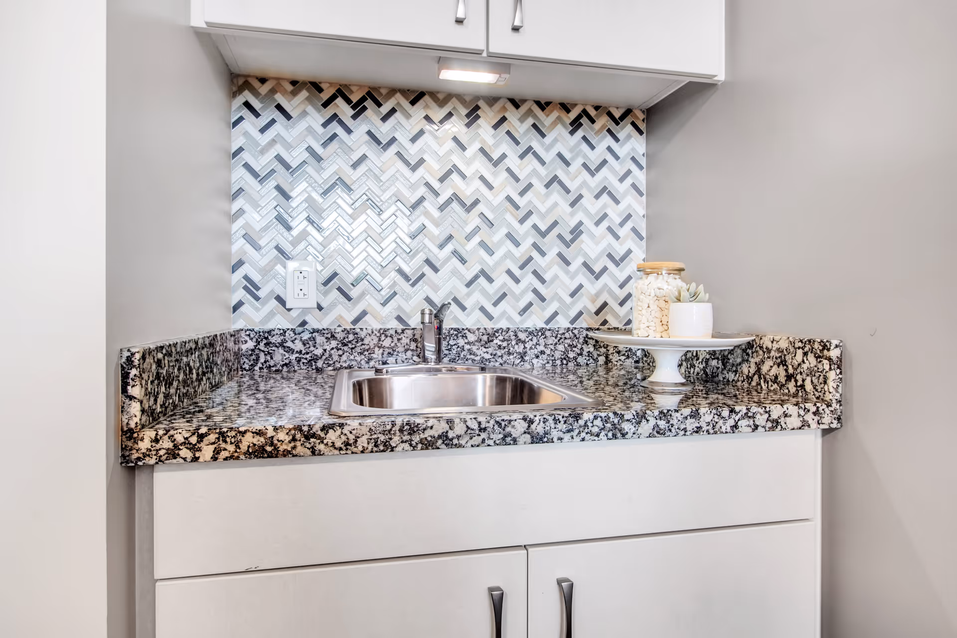 Compact kitchenette with a stainless steel sink, speckled granite countertop, chevron tile backsplash and decorative jar on a pedestal.