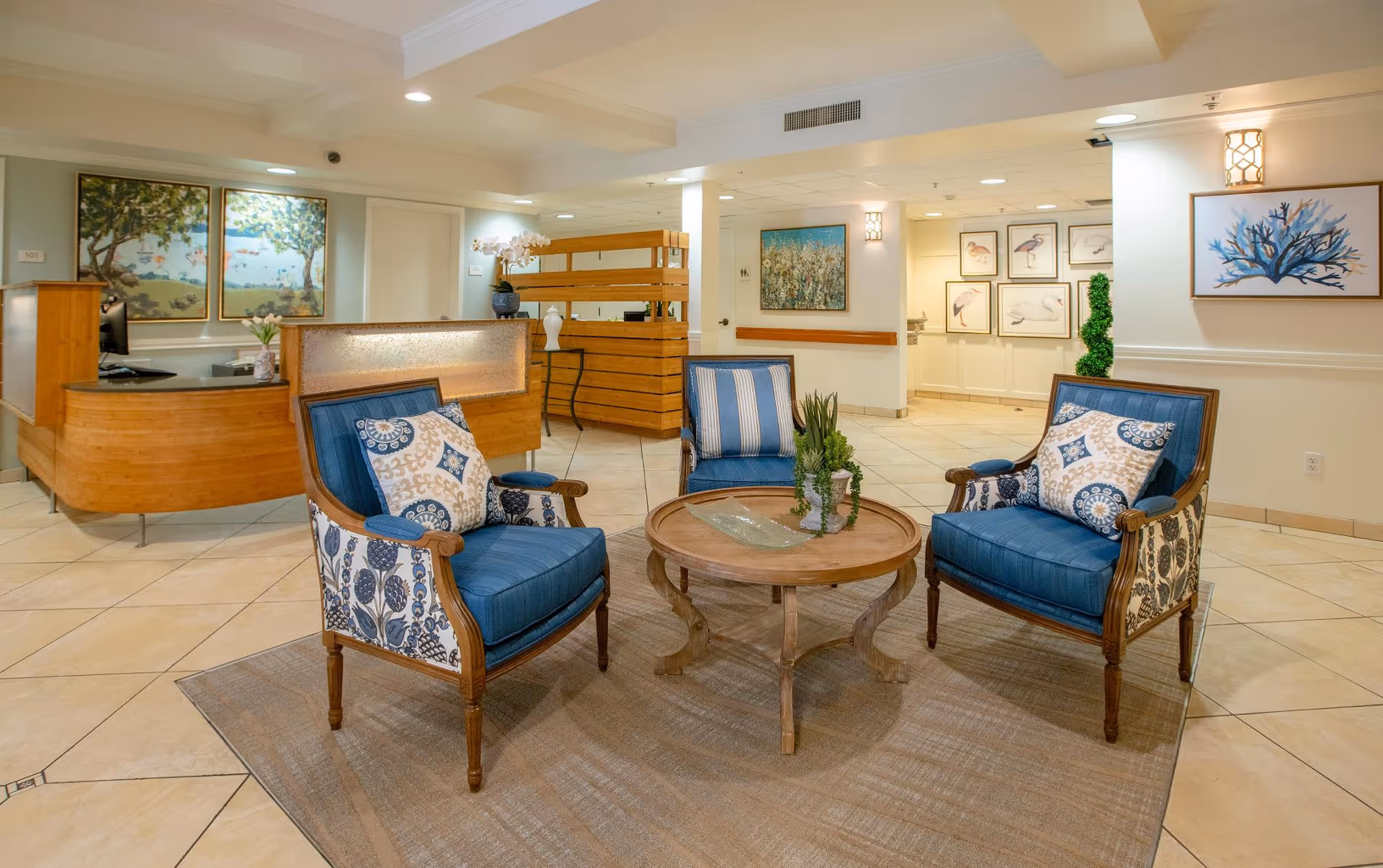 A cozy seating area in a senior living facility lobby with three blue cushioned armchairs arranged around a round wooden coffee table on a beige rug. The background features a wooden reception desk, decorative paintings on the walls, and tiled flooring under warm lighting.