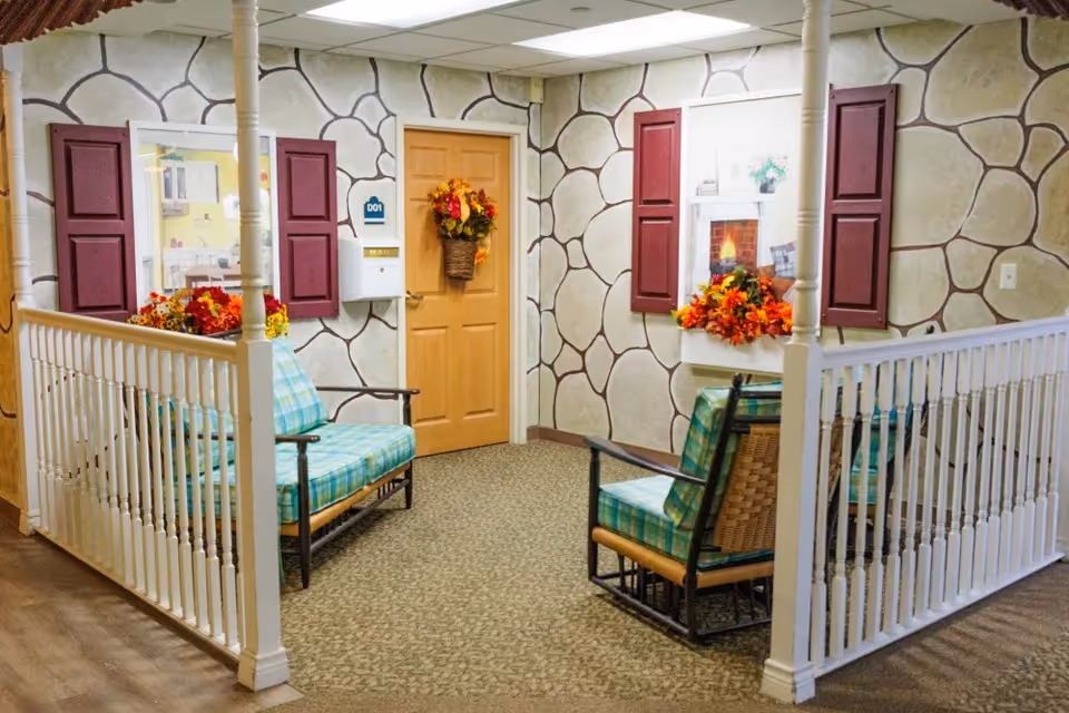 A cozy indoor seating area with two cushioned chairs featuring green plaid upholstery, surrounded by white railings and a wall designed to look like stone with burgundy window shutters. The wall also has two decorative window frames with flower arrangements and a door adorned with a fall-themed wreath.