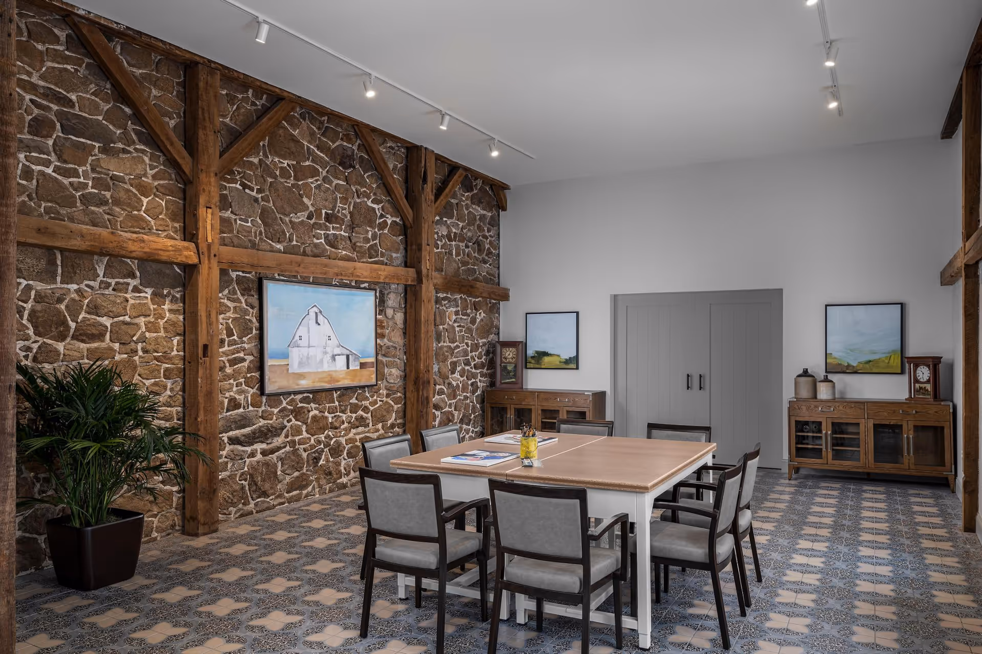 A meeting or dining room with a large square table surrounded by eight chairs. The room features a rustic stone wall with wooden beams, two wooden cabinets with decorative items and framed artwork above them, and a potted plant in the corner. The floor has a patterned tile design, and the ceiling has track lighting.