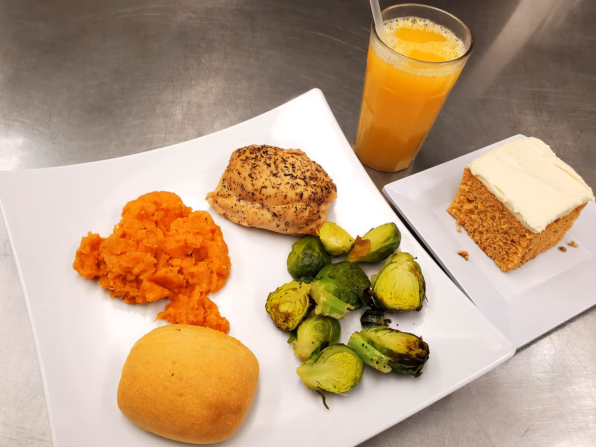 A meal served on a white square plate consisting of mashed sweet potatoes, a seasoned baked chicken thigh, roasted Brussels sprouts, and a dinner roll. Next to the plate is a glass of orange juice with a straw and a square white plate holding a piece of frosted cake.