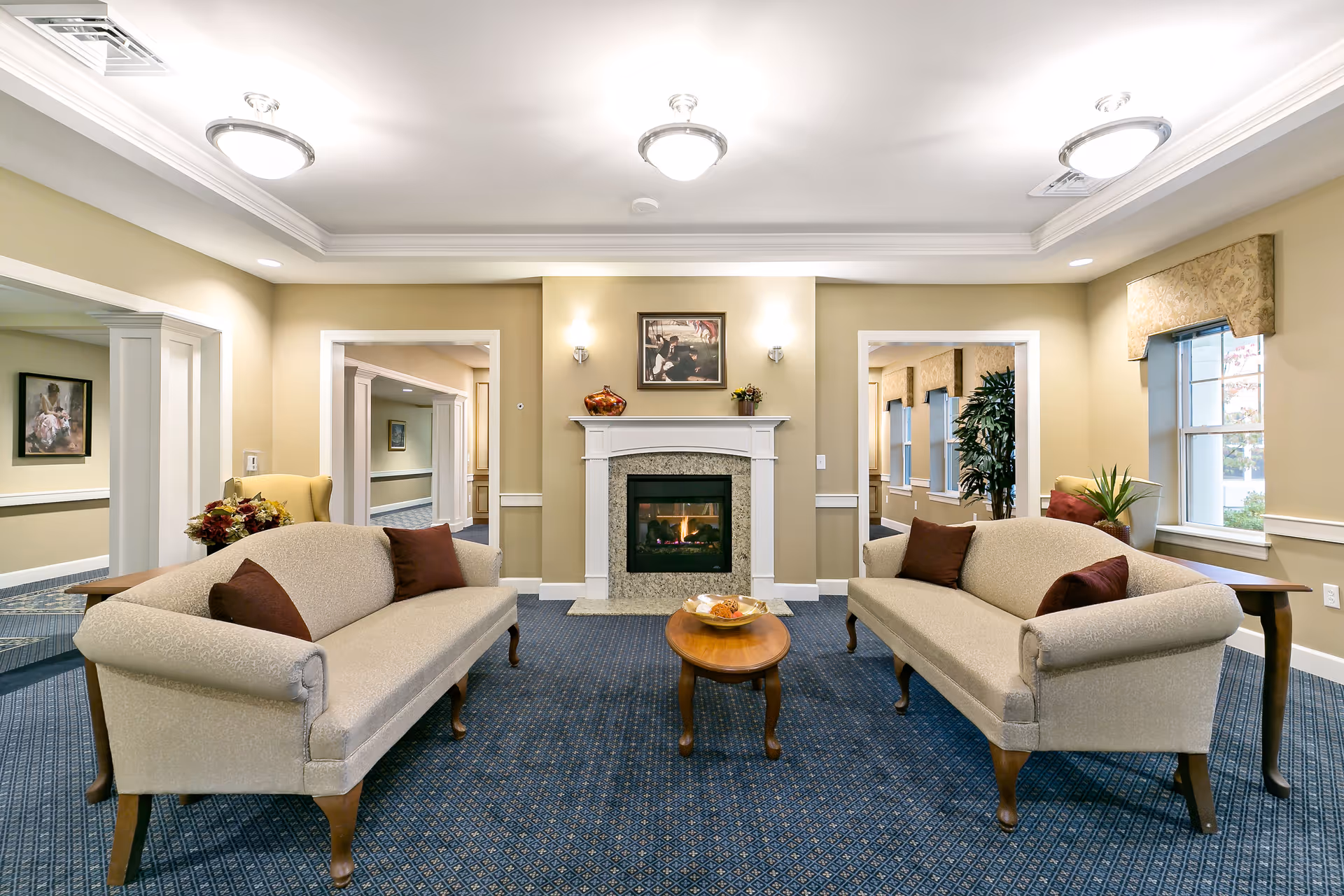 A cozy living room area in an assisted living facility featuring two beige upholstered sofas with brown cushions facing each other across a small wooden coffee table. The room has a blue patterned carpet, beige walls, and a white fireplace with a fire burning inside. Above the fireplace is a framed painting, and there are wall sconces on either side. The room is well-lit with ceiling lights and natural light coming through windows with beige valances. There are doorways leading to other parts of the facility and decorative plants and flowers placed around the room.