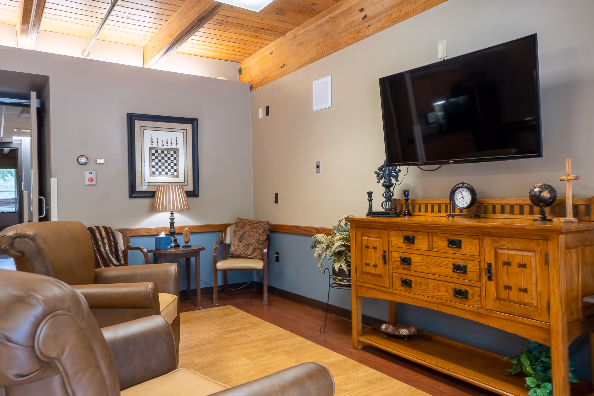 A cozy living room area with two brown leather armchairs, a wooden side table with a lamp and tissue box, a wooden chair with a decorative pillow, and a wooden cabinet with drawers and decorative items including a clock, globe, and cross. A flat-screen TV is mounted on the wall above the cabinet. The room has a wooden ceiling with exposed beams and a window near the ceiling letting in natural light.