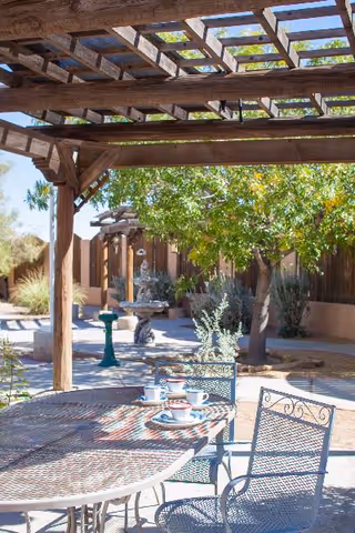 Outdoor patio area with a wooden pergola providing partial shade over a metal table and chairs. The table has several cups and saucers arranged on it. In the background, there is a garden with trees, shrubs, a stone fountain, and a wooden fence under a clear blue sky.