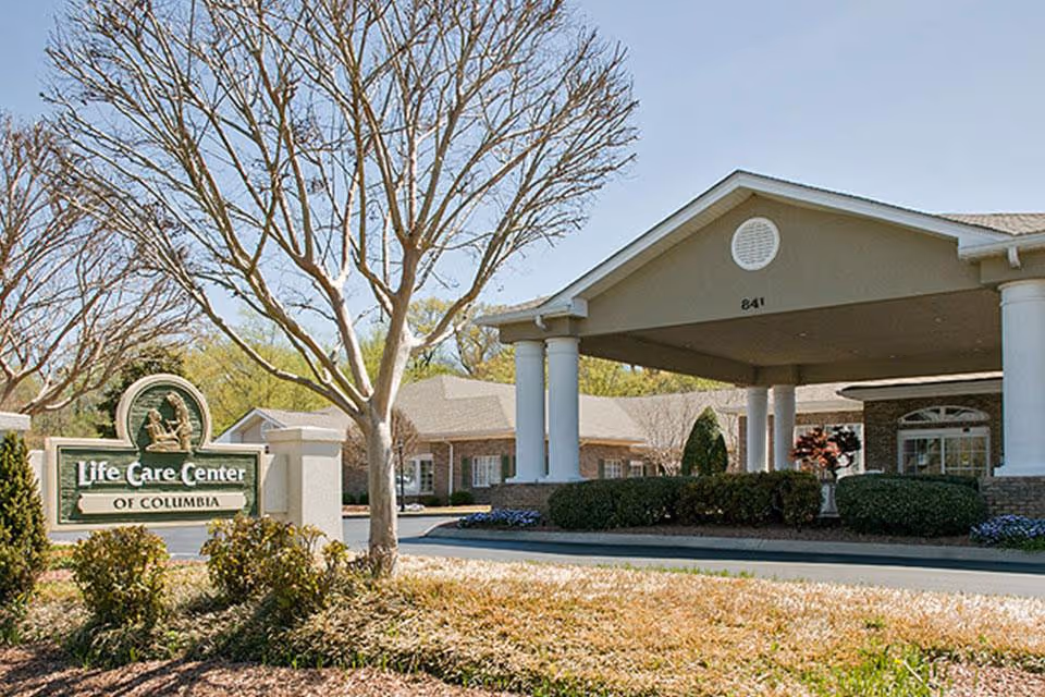 Exterior view of Life Care Center of Columbia, Tennessee showing the building entrance with a covered driveway supported by white columns, surrounded by bushes and trees with no leaves, under a clear blue sky.
