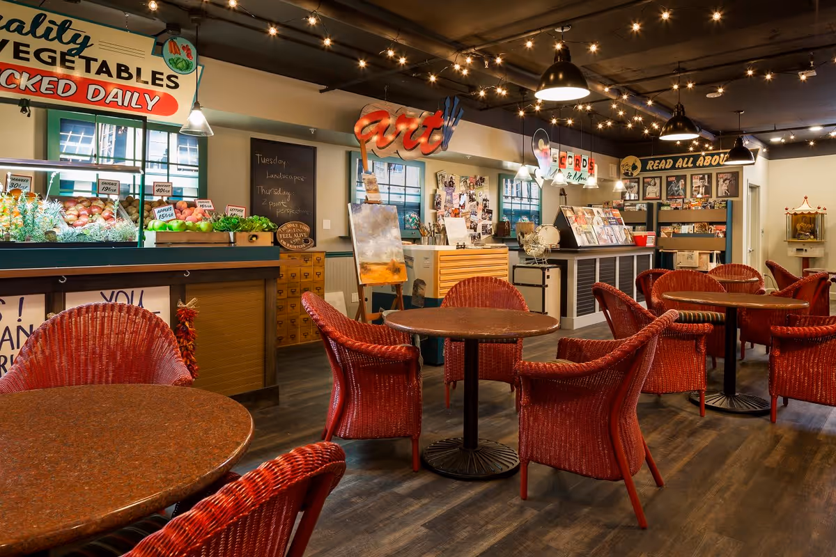 A cozy indoor space with round tables and red wicker chairs arranged for seating. The room features a produce stand with fresh vegetables and fruits, an art display area with paintings and supplies, and a reading section with magazines and books. Warm lighting and decorative signs add to the inviting atmosphere.