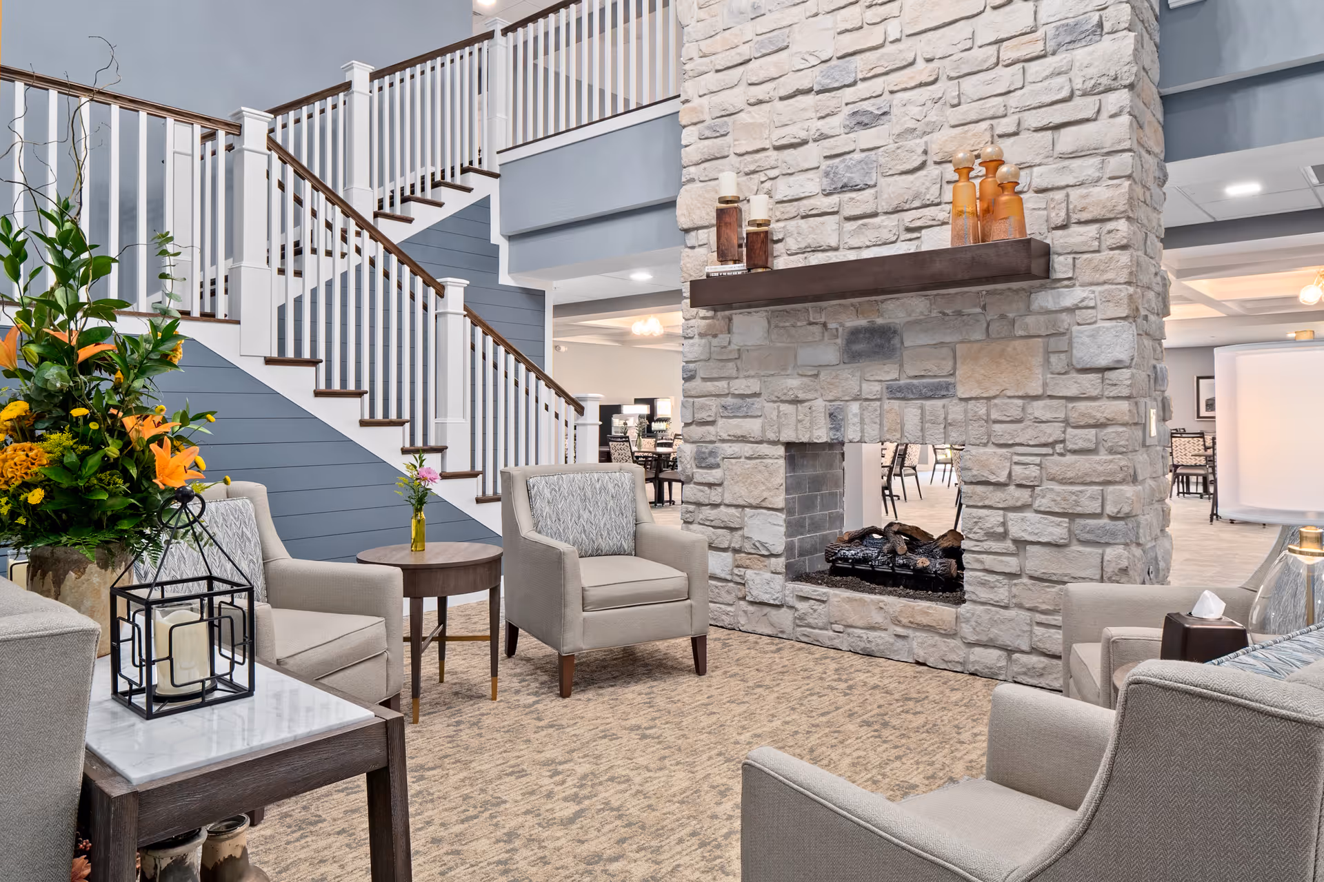 Spacious senior living common area with a stone double-sided fireplace, comfortable armchairs, floral arrangements, and a staircase.