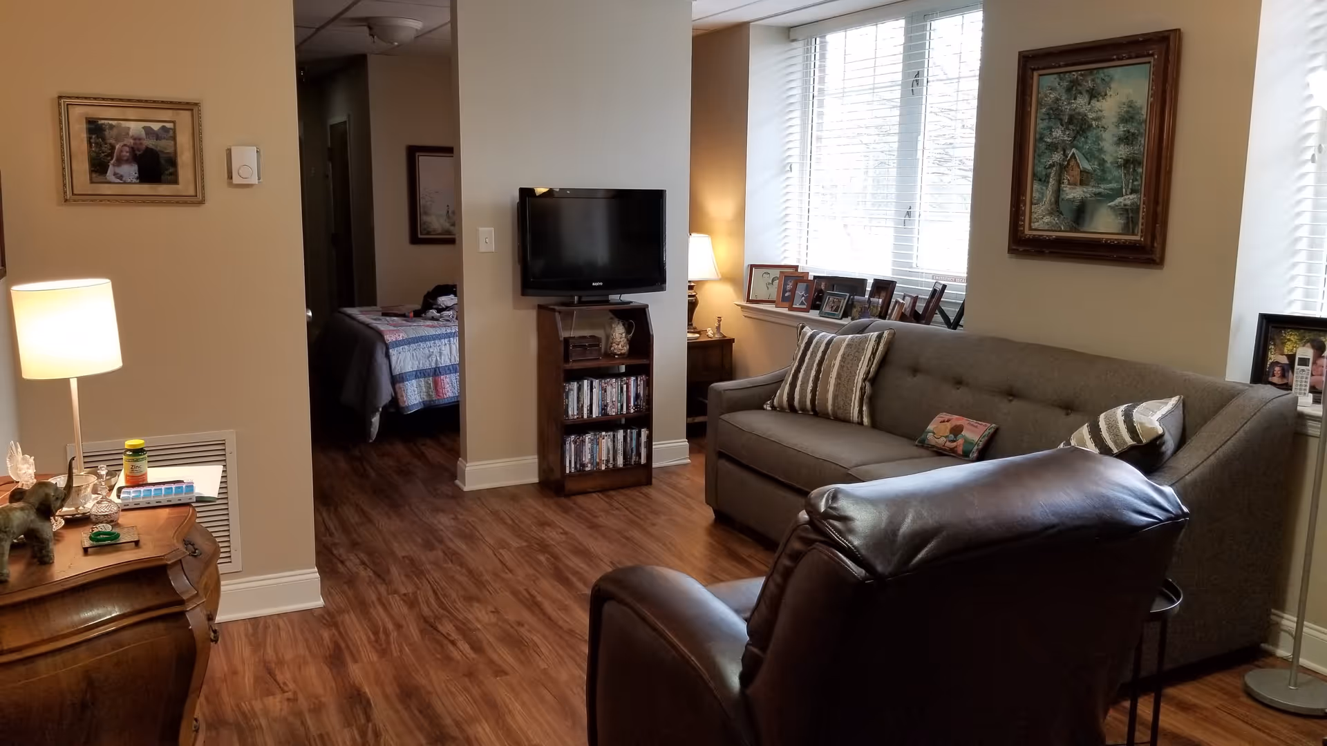 A cozy living room area with a gray sofa adorned with striped and decorative pillows, a dark leather armchair, and a small wooden TV stand holding a flat-screen TV and DVDs. There is a wooden side table with a lamp, medication bottles, and decorative items. The room has wood flooring and beige walls with framed pictures. A bedroom with a bed covered in a quilt is visible through an open doorway.