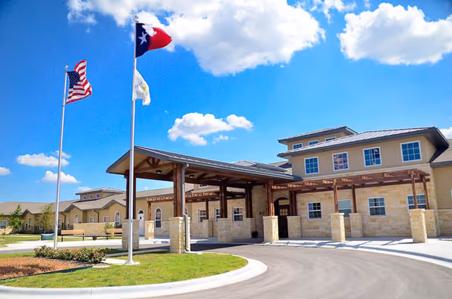 Exterior view of a senior living facility building with a covered entrance, stone pillars, and three flagpoles displaying the American flag, Texas state flag, and another flag. The sky is blue with some clouds.