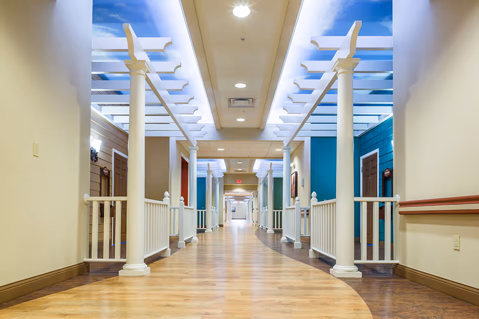 A brightly lit indoor hallway with wooden flooring and white columns supporting a pergola-style ceiling. The walls are painted in neutral and teal colors, with doors along the sides. The ceiling has recessed lighting and a section with a sky mural featuring clouds.
