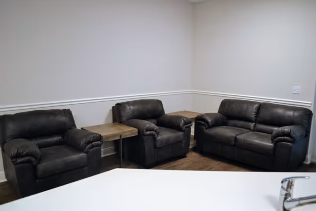 A small seating area with two black leather armchairs and one black leather loveseat arranged around two wooden side tables in a corner of a room with white walls and wood flooring. A white countertop with a faucet is visible in the foreground.