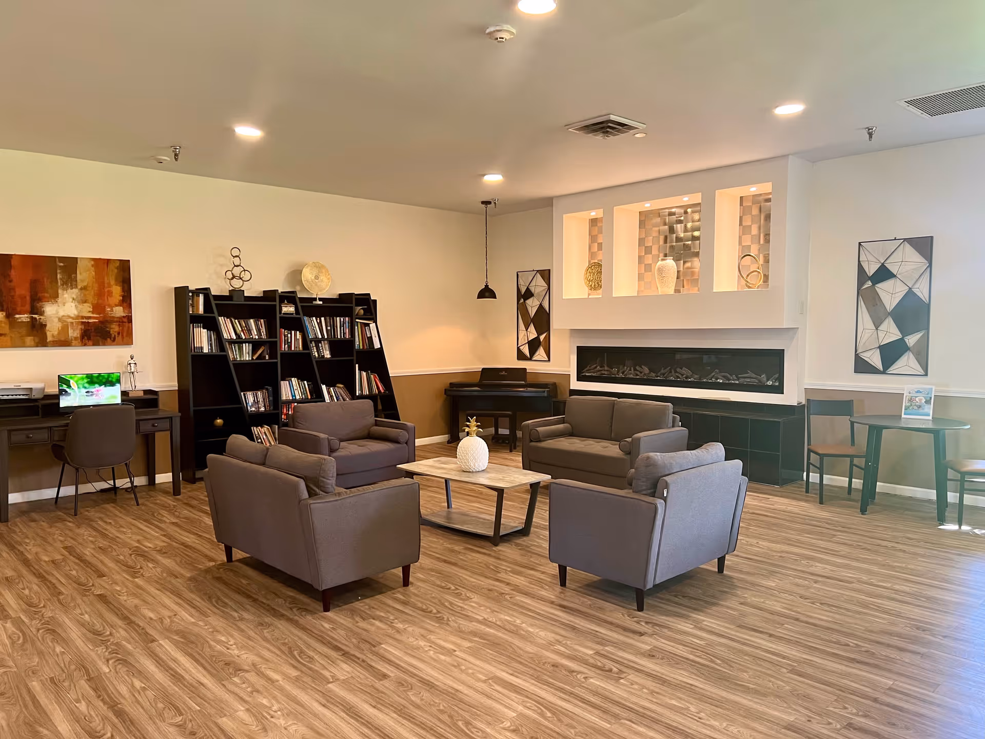 A cozy senior living common area with four gray armchairs arranged around a wooden coffee table with a decorative pineapple. The room features wood flooring, a built-in modern fireplace, a piano in the corner, bookshelves filled with books, a desk with a chair and a computer, and decorative wall art.