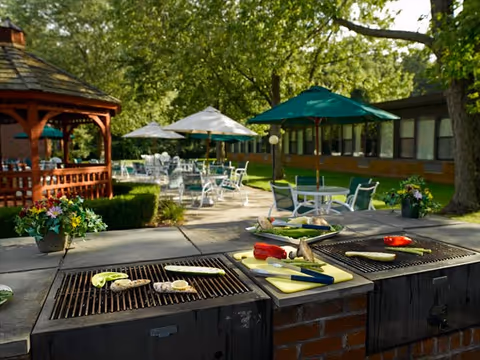 Outdoor barbecue grills with vegetables like zucchini, mushrooms, and red peppers being cooked. In the background, there are patio tables with umbrellas and chairs set in a garden area with trees and a wooden gazebo.