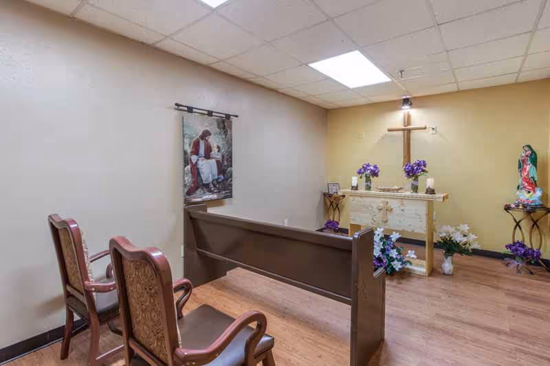 A small chapel room with wooden pews and chairs facing an altar. The altar has a wooden cross mounted on the wall above it, with purple flowers and candles placed on and around the altar. A religious painting hangs on the left wall, and a statue of the Virgin Mary is on a small table to the right. The room has a light-colored ceiling with recessed lighting and wood flooring.