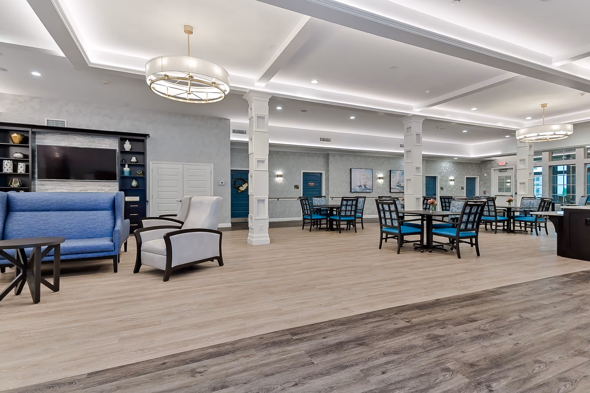 A spacious and modern common area in a senior living facility featuring a seating area with blue and white armchairs, a large flat-screen TV mounted on a wall unit with decorative items, multiple dining tables with blue cushioned chairs, and elegant ceiling lighting fixtures. The room has light wood flooring and soft gray walls with decorative columns.