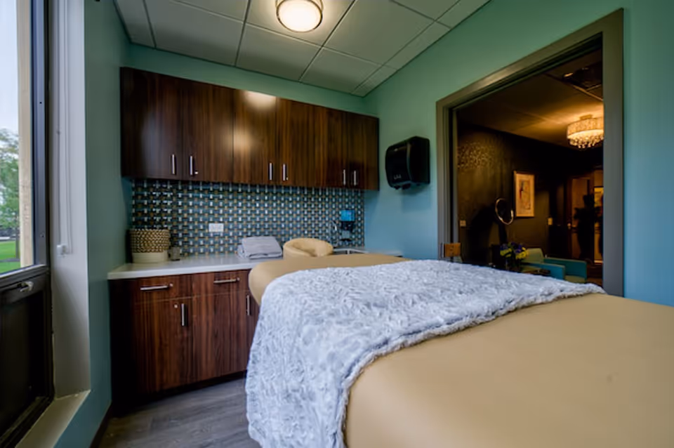 A cozy room with a massage or therapy table covered with a soft gray blanket. The room has wooden cabinets with a patterned backsplash, a paper towel dispenser, and a window letting in natural light. An adjacent room with warm lighting and a chandelier is visible through an open doorway.