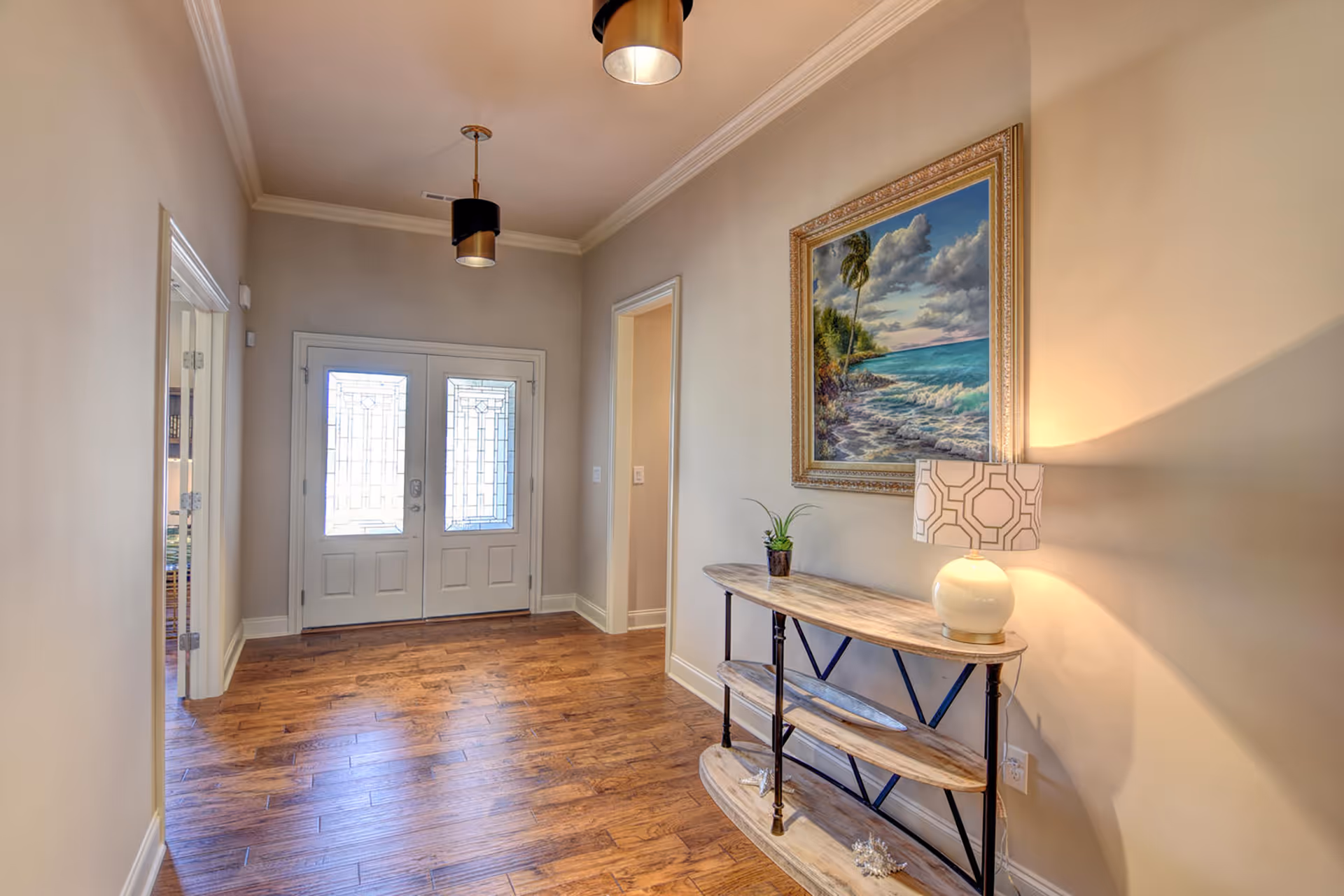 A bright entryway with double glass-paneled doors, wooden flooring, and beige walls. A wooden console table with three shelves is placed against the right wall, holding a small potted plant and a lamp with a patterned shade. Above the table hangs a framed painting of a tropical beach scene with palm trees and ocean waves. Two ceiling light fixtures with black and gold accents illuminate the space.