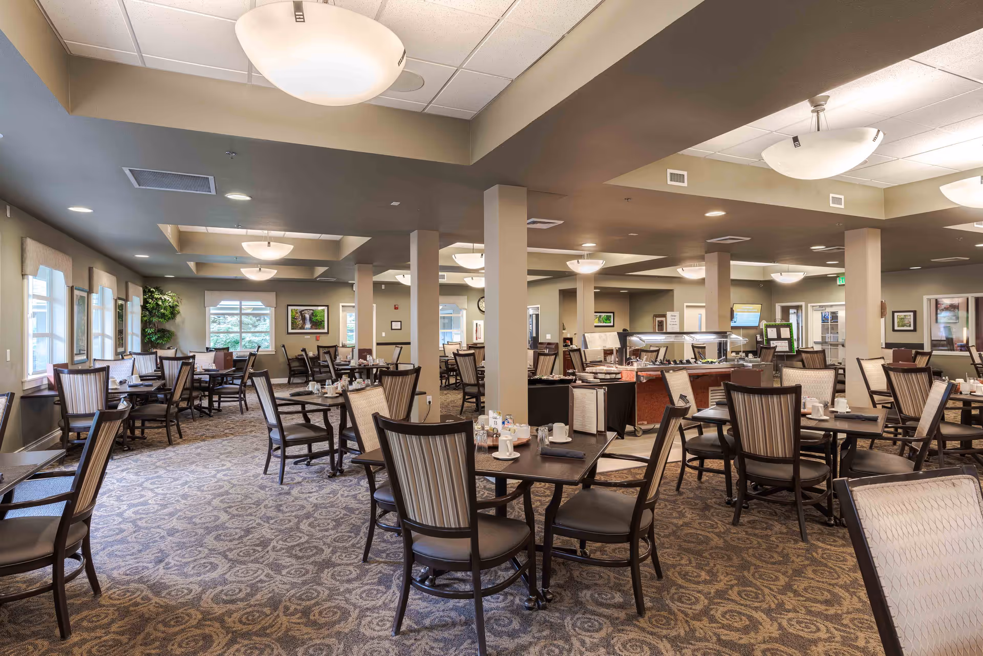 A spacious dining room in a retirement community with multiple tables and chairs arranged neatly. The room features patterned carpet flooring, large windows with curtains, ceiling lights, and a buffet serving area in the center. The walls are decorated with framed pictures and there are several columns supporting the ceiling.