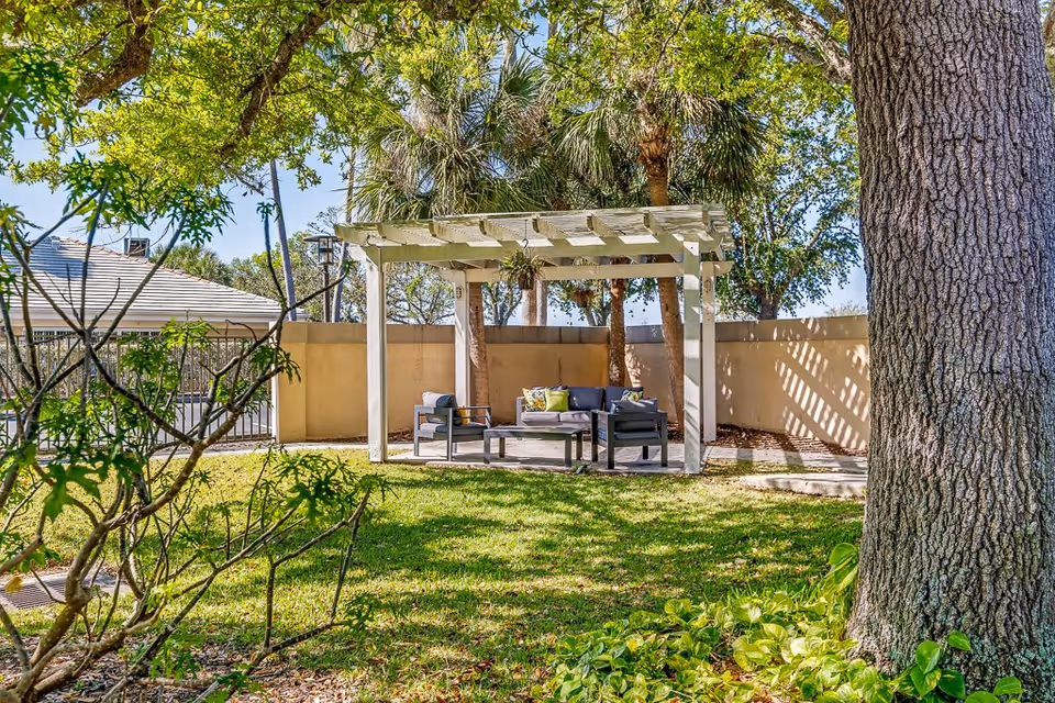 Outdoor garden area with green grass, trees, and a white pergola structure. Under the pergola, there is outdoor seating including a sofa and two chairs with cushions, and a coffee table. The area is surrounded by a beige wall and there are palm trees and other greenery in the background.