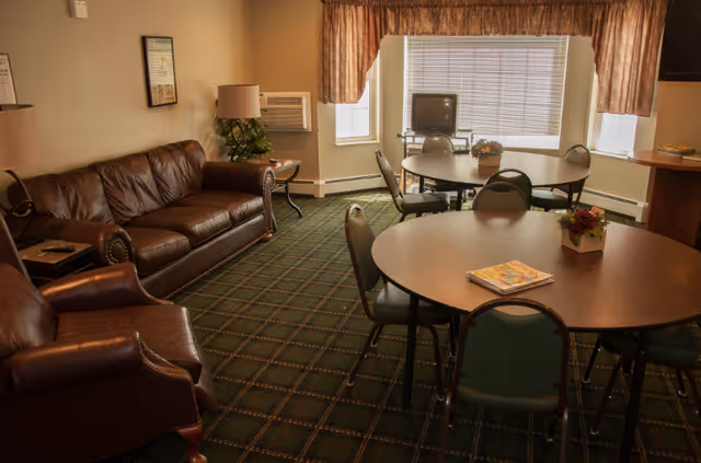 A senior living common room with round tables and chairs, leather sofas, a small TV, and a large window with curtains.
