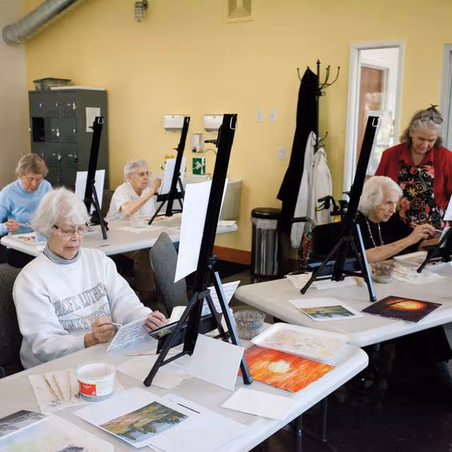 A group of elderly women seated at tables in a room, engaged in painting on canvases set on easels. Art supplies and paintings are visible on the tables. A woman stands assisting one of the seated women.