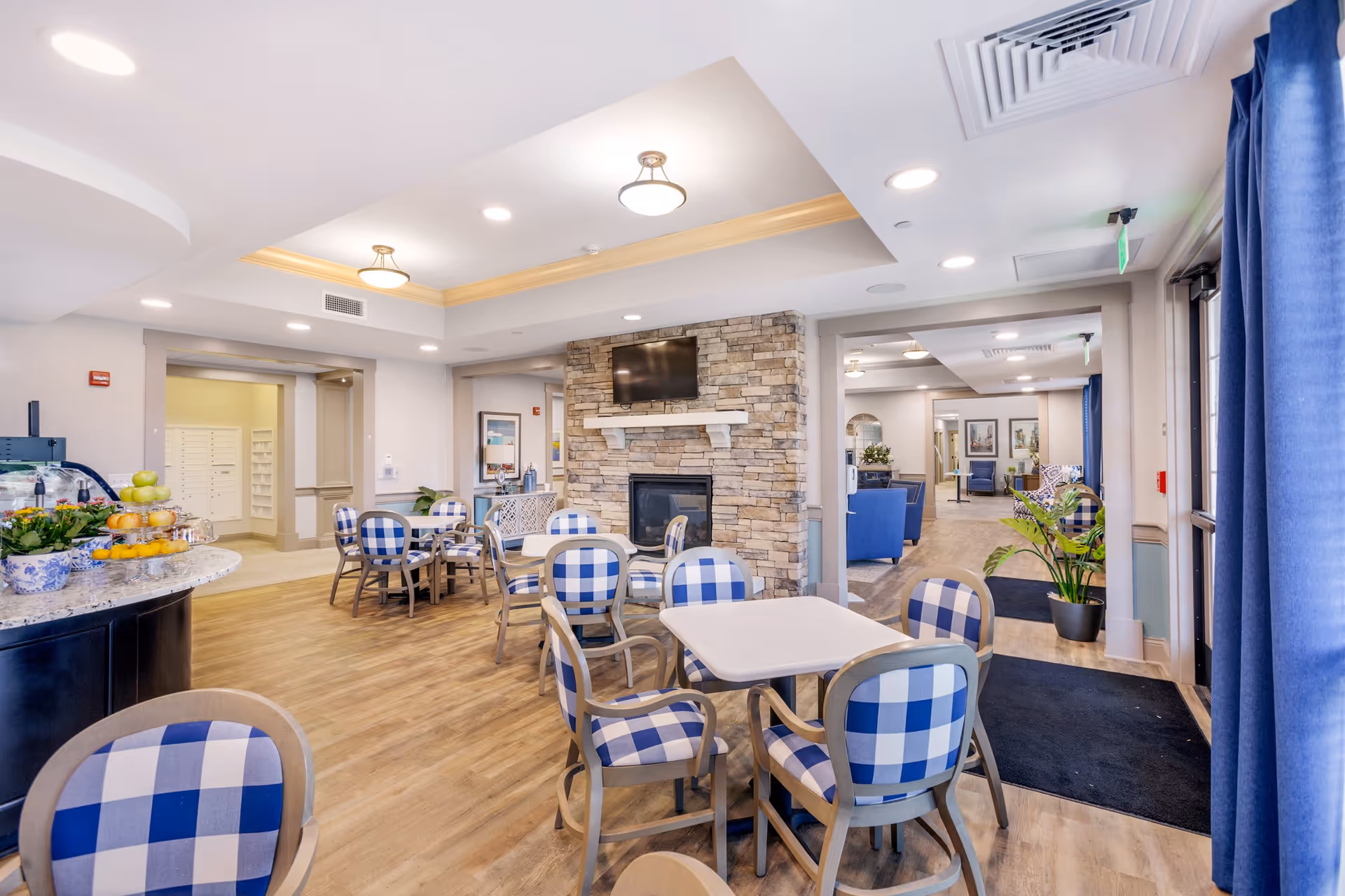 A bright and spacious common area in a senior living facility featuring several tables with blue and white checkered chairs. The room has wood flooring, a stone fireplace with a mounted TV above it, and recessed lighting in the ceiling. There is a counter with fruit and plants on the left side and a hallway leading to other rooms in the background. Blue curtains frame a glass door on the right side.