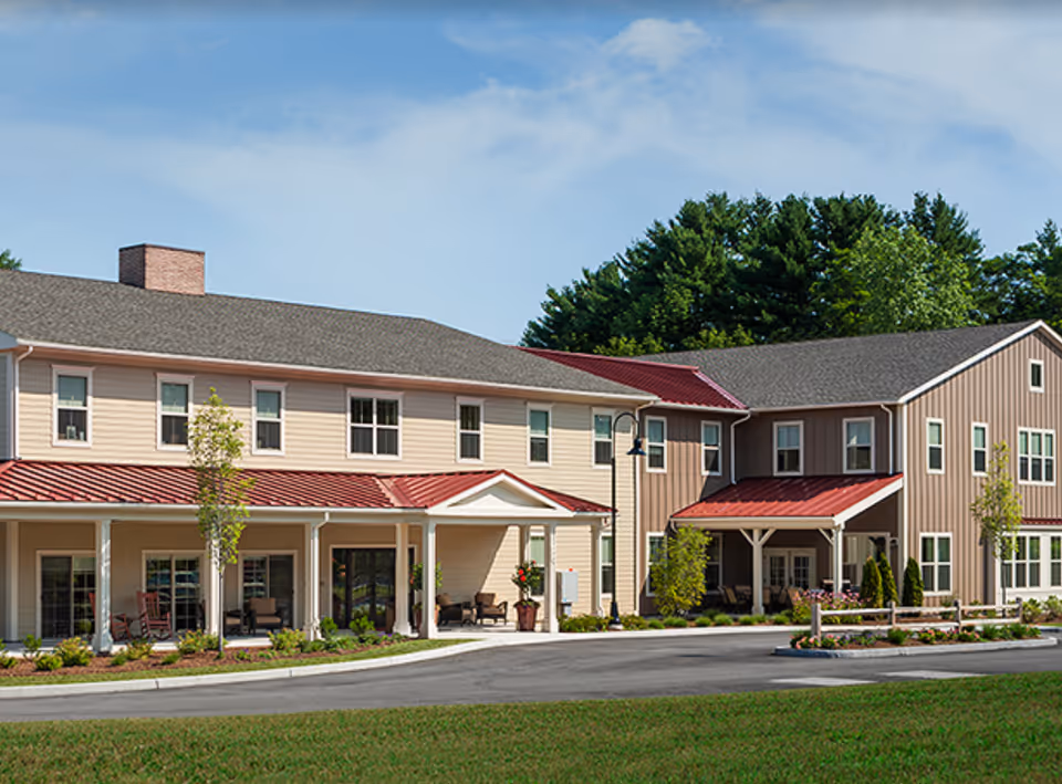 Front exterior of a two-story senior living building with covered porches, red metal awnings, and a landscaped entrance.