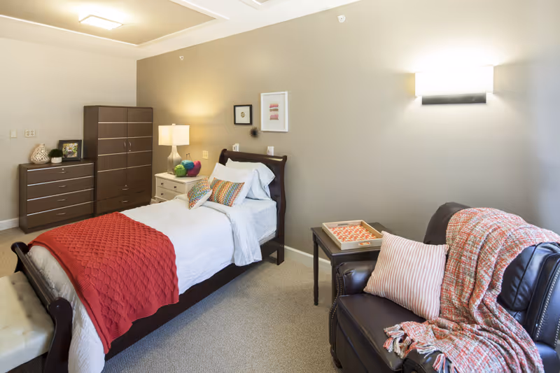 A cozy bedroom in a senior living facility featuring a single bed with white bedding and a red throw blanket, colorful pillows, a wooden dresser and wardrobe, a nightstand with a lamp, and a dark leather armchair with a striped pillow and a red patterned throw blanket. The walls are painted beige with framed artwork and a wall-mounted light fixture.