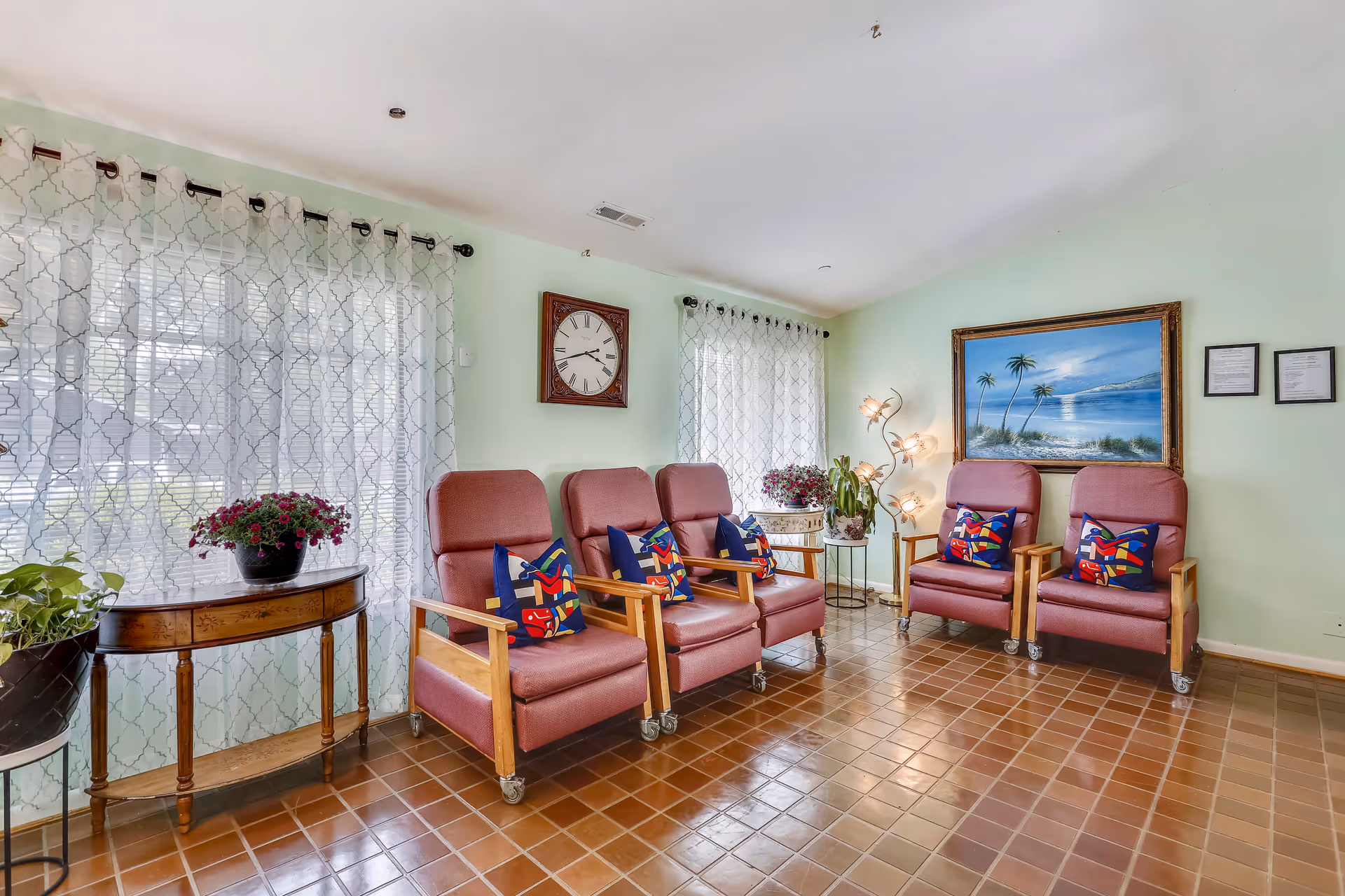 A cozy sitting area with five cushioned chairs with wooden armrests and wheels, each adorned with colorful patterned pillows. The room has light green walls, two large windows with sheer patterned curtains, a wall clock, a large framed painting of a beach scene with palm trees, and two framed documents. There are also several potted plants on a wooden table and stands, and a unique floor lamp with multiple light fixtures. The floor is covered with brown square tiles.
