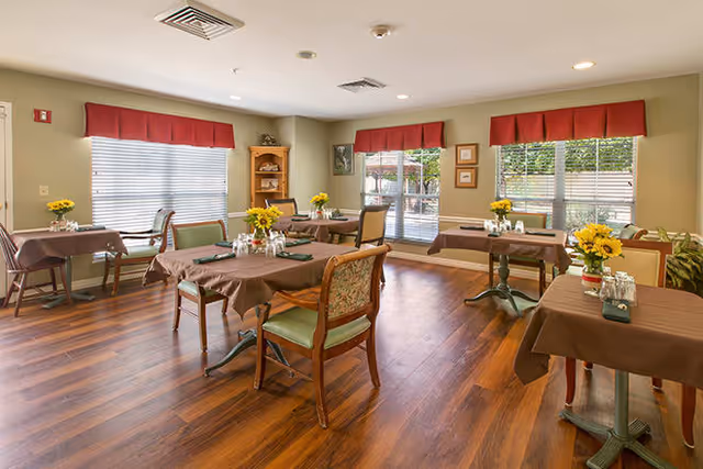 A dining room with several tables covered in brown tablecloths, each set with napkins, salt and pepper shakers, and a vase of yellow flowers. The room has large windows with red valances, wooden flooring, and a small wooden shelf in the corner.