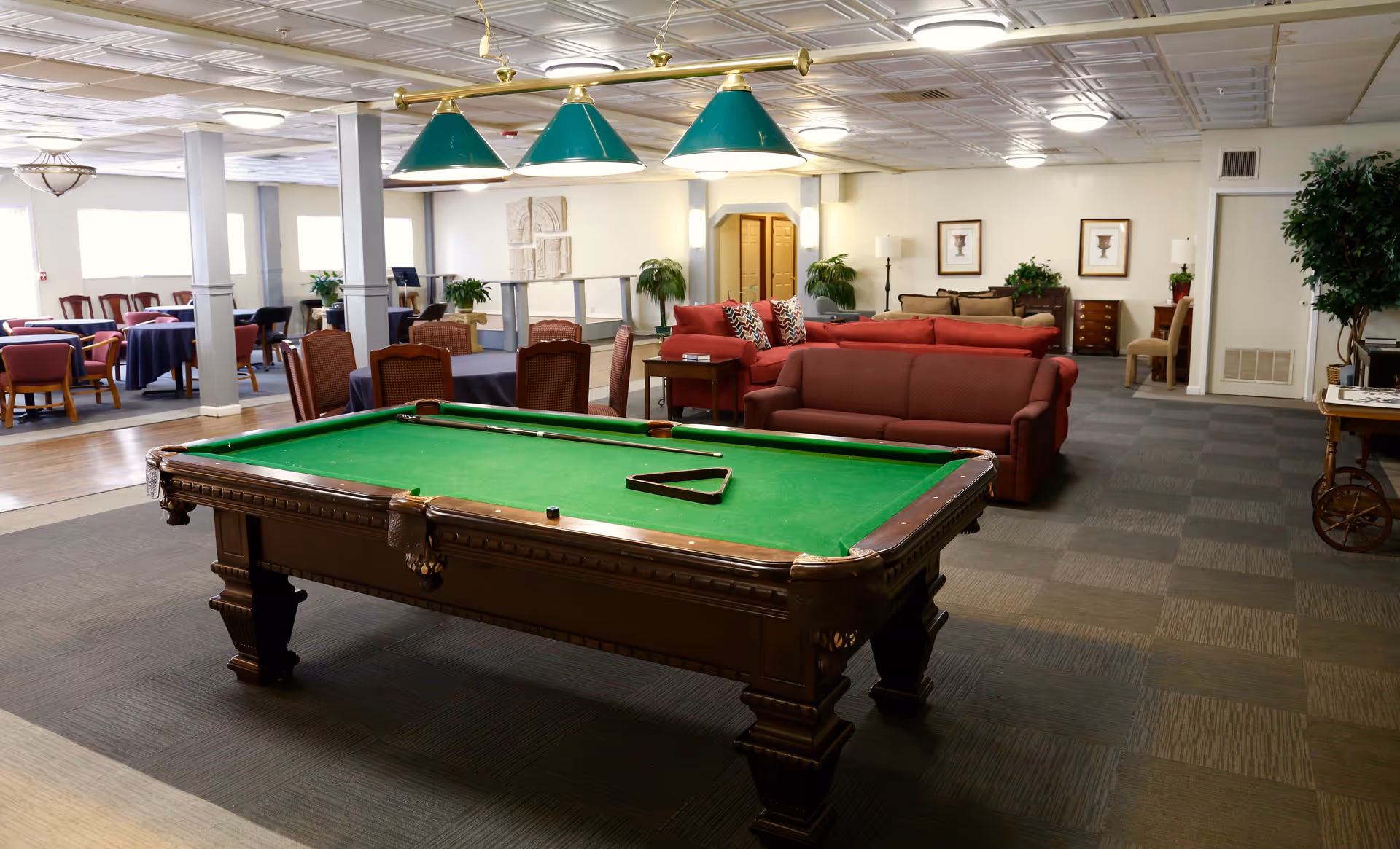 A spacious common area in a senior living facility featuring a green felt pool table with cues and a triangle rack, surrounded by comfortable red sofas and armchairs. In the background, there are several dining tables with chairs, potted plants, framed artwork on the walls, and soft overhead lighting.