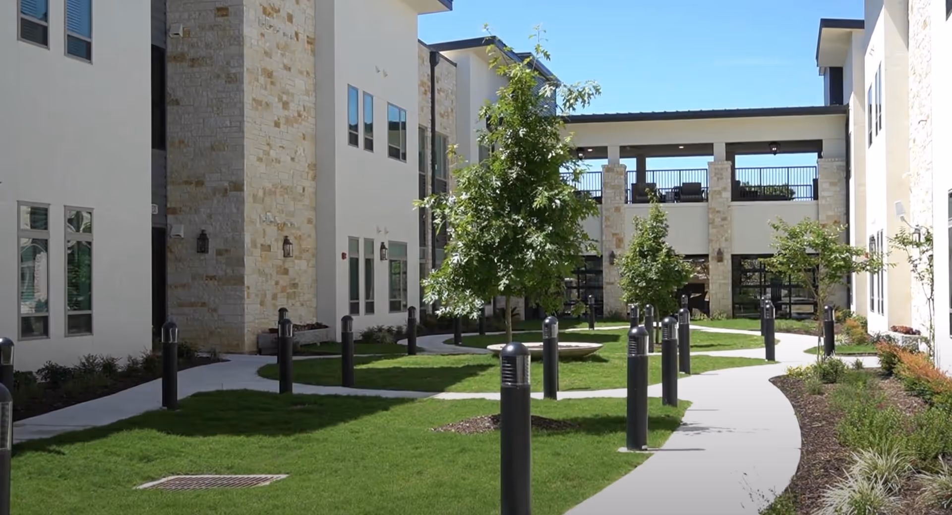 Landscaped courtyard with curved concrete pathways, grass, young trees, bollard lights and surrounding multi-story white stone residential buildings.