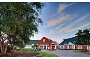 Exterior view of Longwood At Oakmont Retirement Community featuring a red building with white trim, a covered entrance, and a driveway surrounded by trees and landscaping under a partly cloudy sky.
