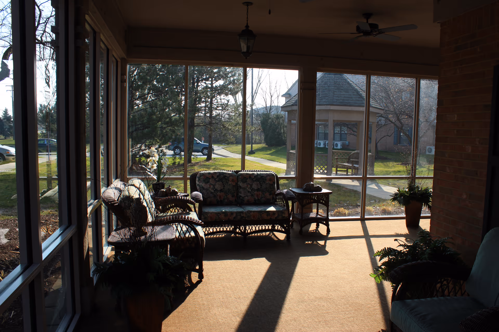 A sunlit enclosed porch area with wicker furniture including a loveseat, two chairs, and a small table. The furniture has floral cushions. Large windows provide a view of a green lawn, trees, and a gazebo outside. There are potted plants inside the porch and ceiling fans above.