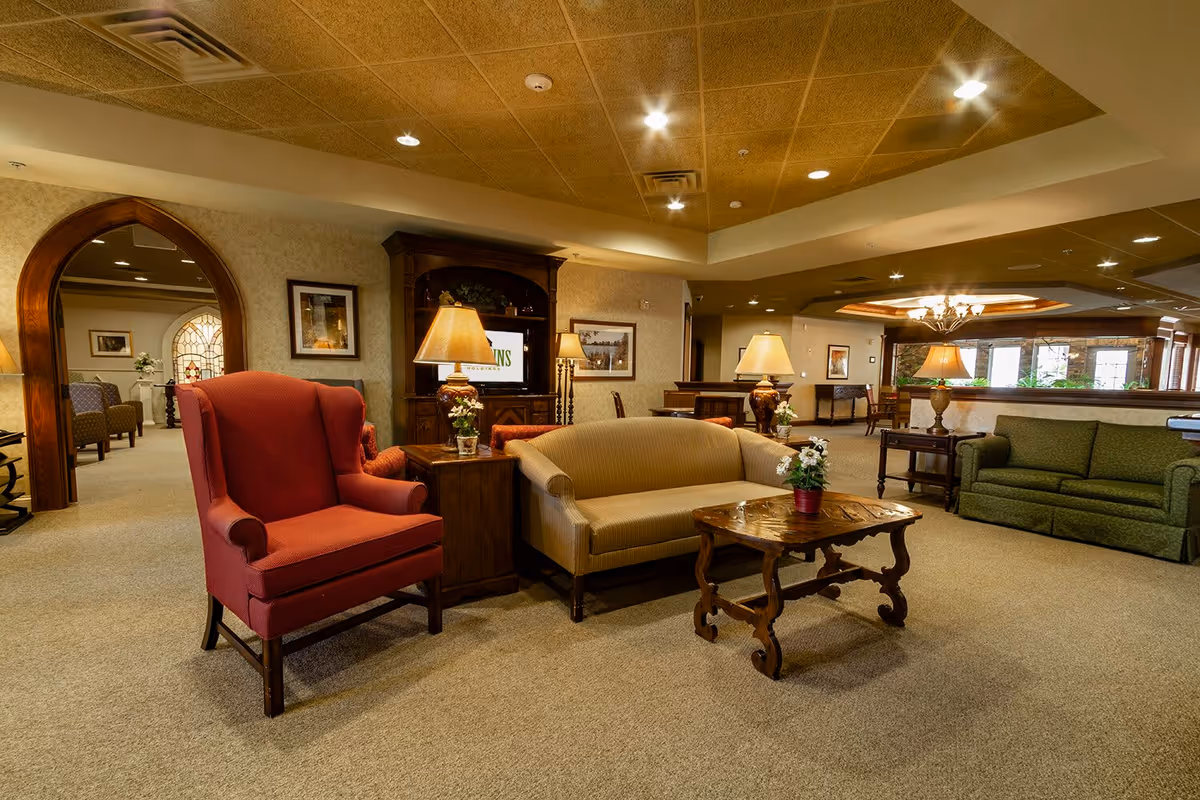 A spacious and warmly lit living room area in a senior living facility featuring a red armchair, a beige sofa, a green sofa, wooden side tables with lamps and small flower pots, a wooden coffee table, and a large wooden cabinet with a television. The room has a carpeted floor, decorative archway, framed pictures on the walls, and recessed lighting in the ceiling.