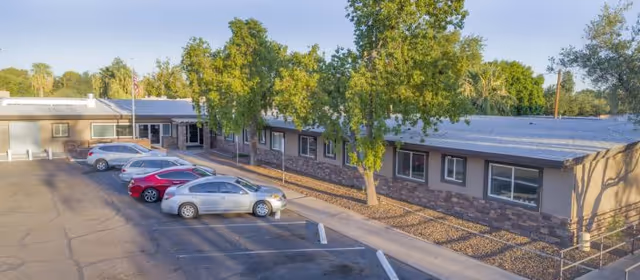 Exterior view of Desert Terrace Healthcare Center showing a single-story building with multiple windows, a parking lot with several parked cars, and trees lining the sidewalk in front of the building under a clear sky.