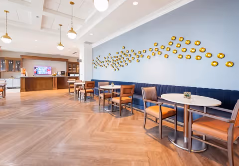 A spacious dining area with wooden flooring, round tables, and chairs with orange cushions. A long blue cushioned bench runs along a light blue wall decorated with a pattern of small golden circular wall art. Pendant lights hang from the ceiling, and a wooden counter with cabinets and a TV is visible in the background.